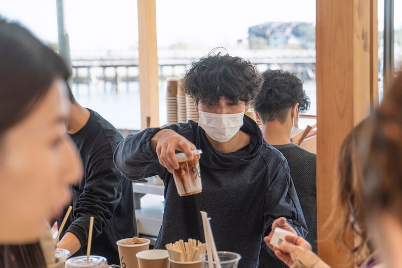 Una persona joven con una sudadera con capucha negra y una mascarilla sostiene una bebida de café helado en una mesa, rodeada de amigos.
