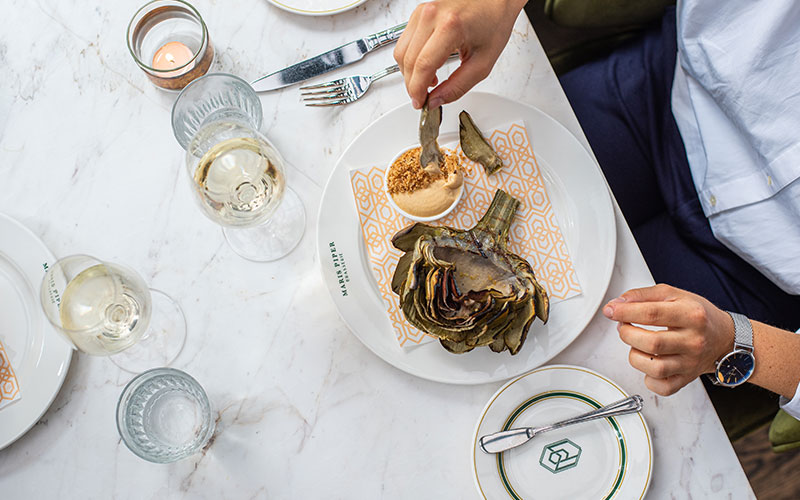 An overhead shot of someone dipping an artichoke leaf into a dip The place setting is of a nice restaurant with a white marble table white and gold plates and wine glasses