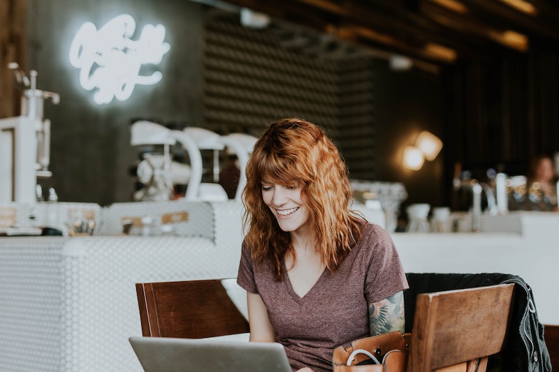 A woman with red hair wearing a mauve colored t shirt sits in a cafe working on her laptop