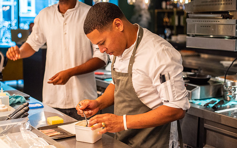 A male chef in a white chef's coat and gray apron stands in a restaurant kitchen spooning something out of a small white container.