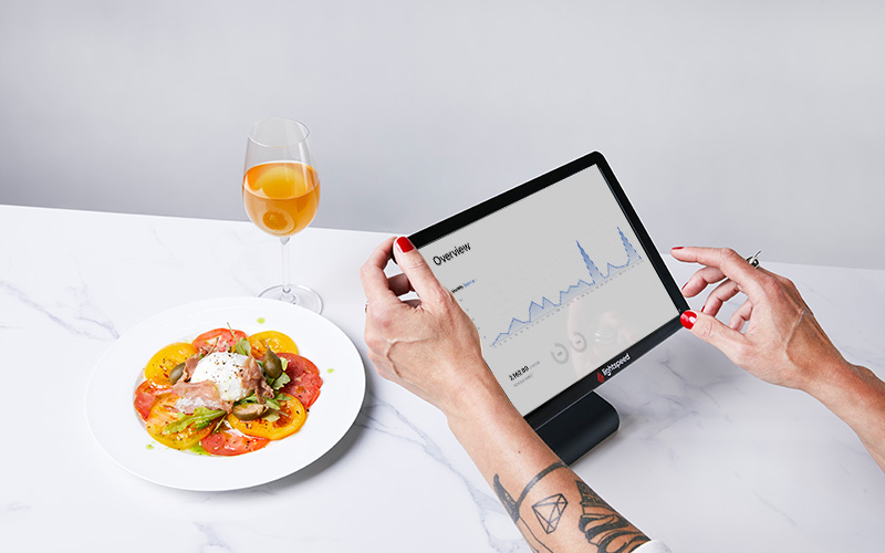 A person's hands using Lightspeed POS to check their reports. A glass of wine and plate of tomato salad sit next to them.