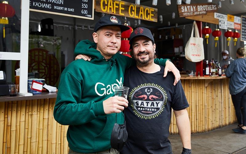 A man in a green hoodie with his arm around a man in a black t-shirt both stand in front of an outdoor cafe, looking at the camera.