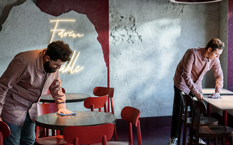 Two bussers in red shirts wipe down tables in a restaurant dining room.