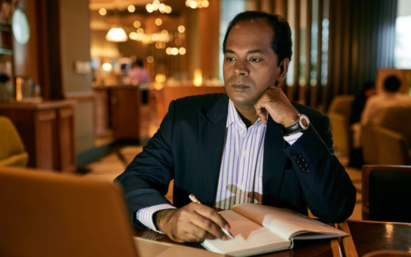 A man in a suit sits in an empty, dimly-lit restaurant. He's looking at his laptop open in font of him while he writes in a notebook.