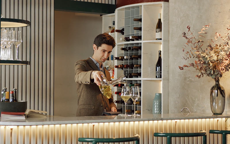 A man in a brown suit stands behind a bar pouring two glasses of white wine.