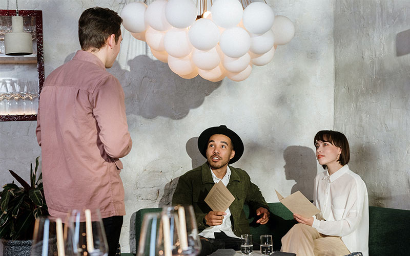 A manager in a red shirt stands with his back to the camera, talking to two restaurant guests sitting at a table.