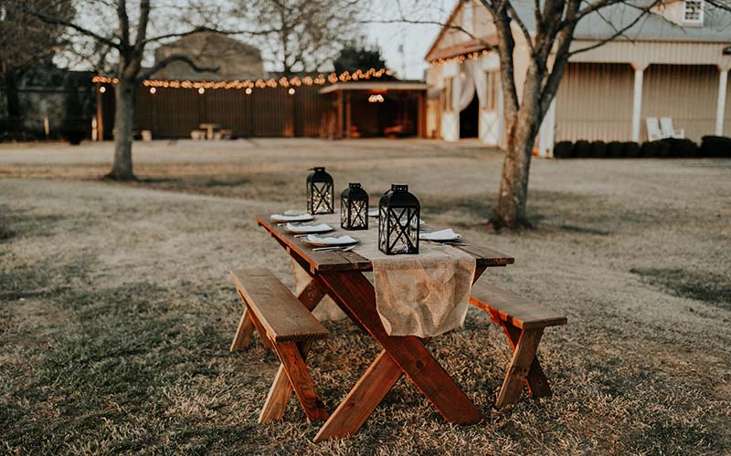 An outdoor picnic table set up with a barn in the background decorated with string lights.