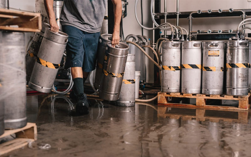 A person wearing blue shorts and a gray shirt carries mini kegs on the brewery floor.