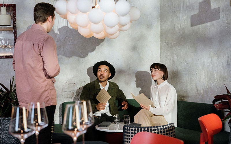 Two people sit at a table with menus in hand, looking up at the server who is speaking to them.