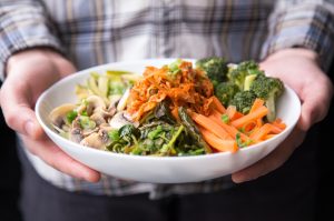 Close up of two hands holding a bowl  with mushrooms, carrots, broccoli, and kimchi.