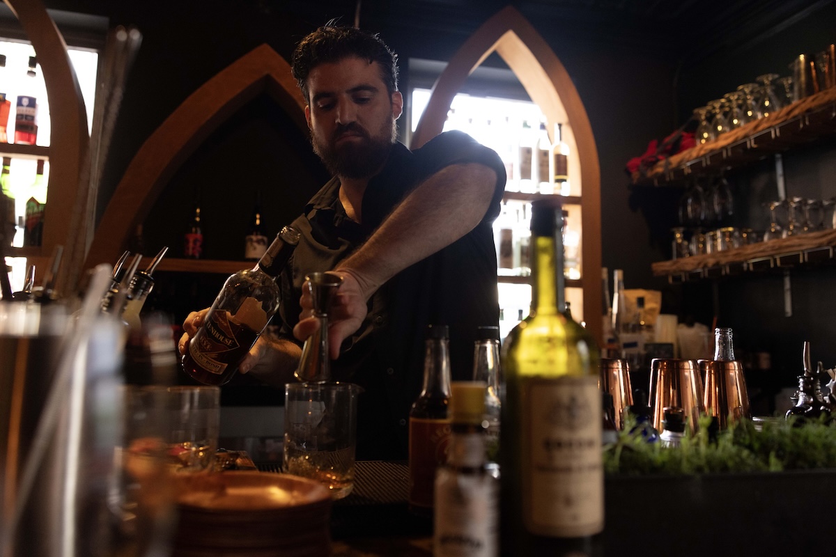  A bartender carefully prepares a drink, pouring liquor into a jigger in a dimly lit bar with shelves of bottles and glasses behind him.