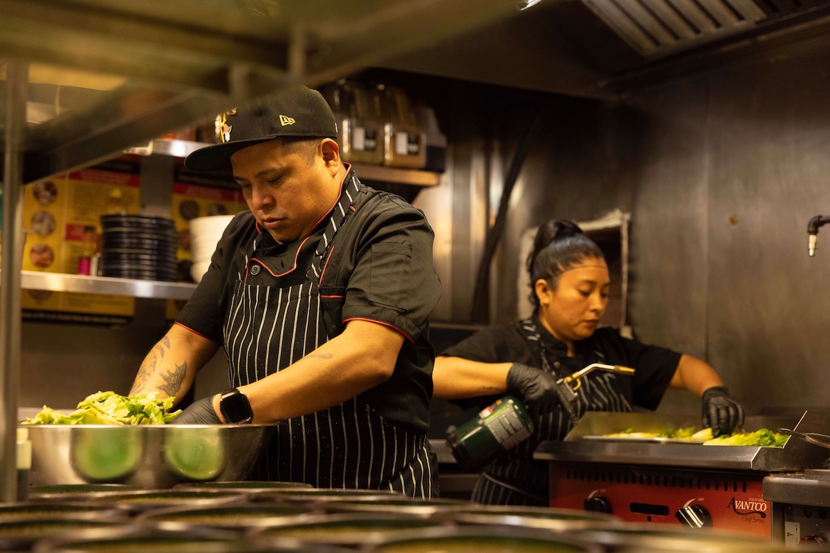 Two chefs, wearing striped aprons and gloves, focus on preparing dishes in a busy commercial kitchen.