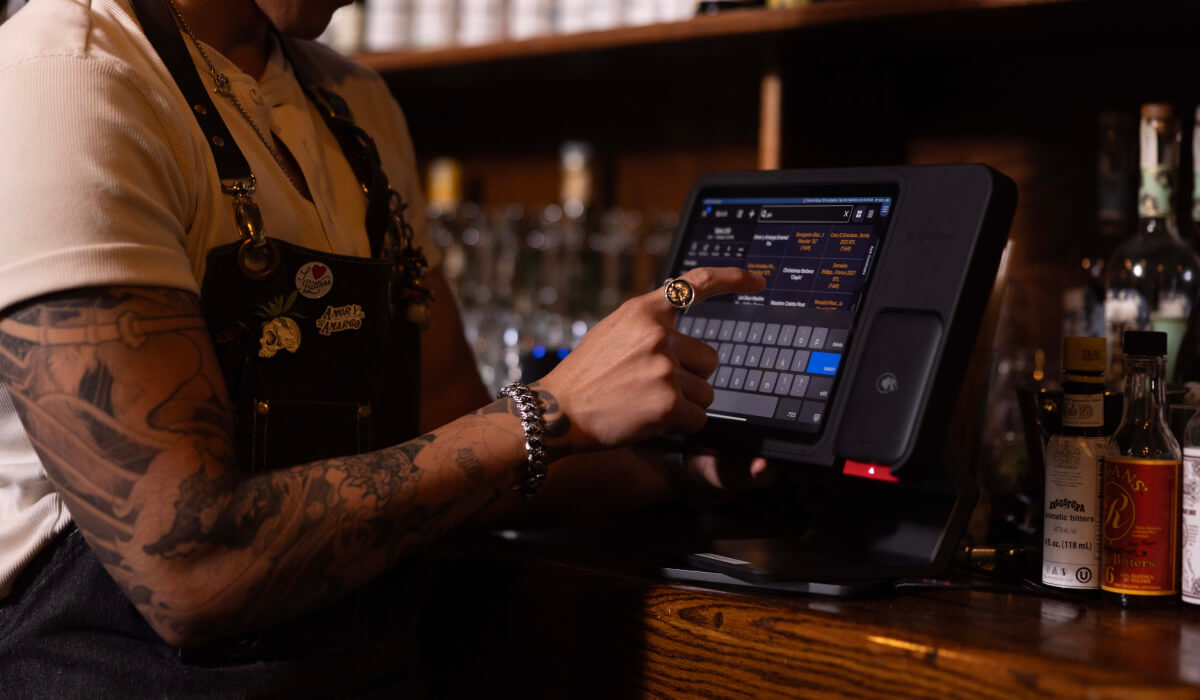 A front of house (FOH) employee uses a touchscreen POS system to input an order in a dimly lit bar, surrounded by bottles on shelves in the background.