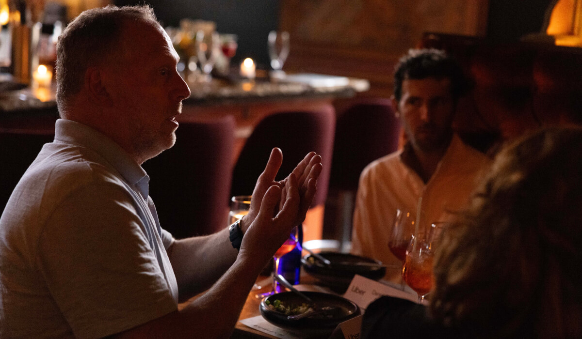 A man engages in a thoughtful discussion at a dimly lit restaurant table, gesturing as he speaks while others listen.